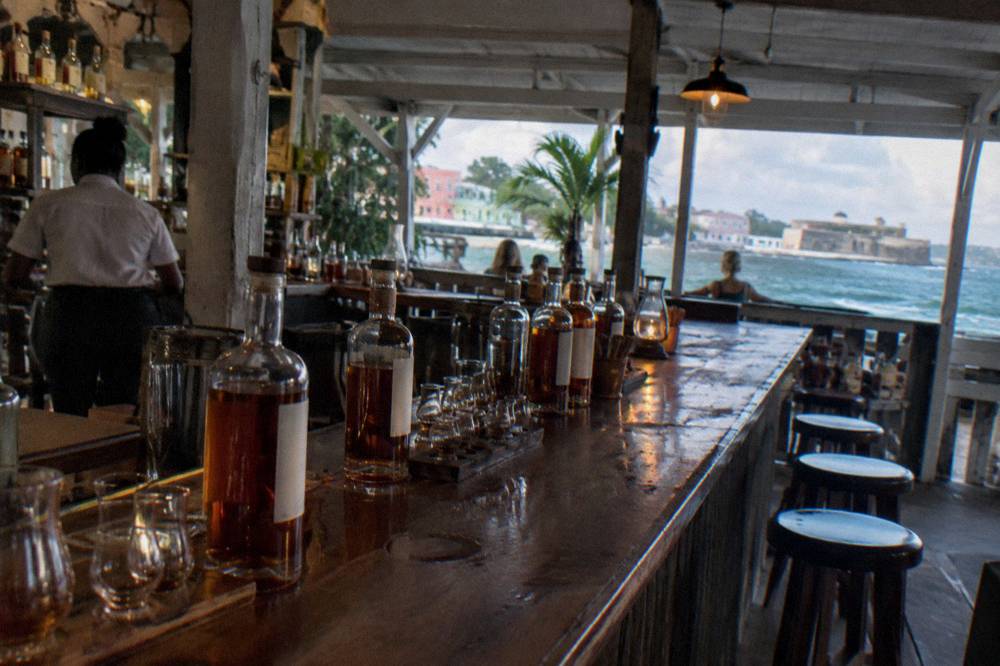 Wooden bar counter lined with rum bottles and tasting glasses in a dim tavern interior